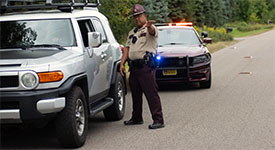 A state trooper next to a vehicle on the side of the road, speaking with the driver through the window.