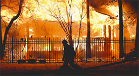 The silhouette of a firefighter walking in front of the flames of a structure fire.