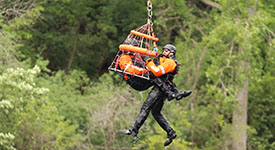 A Minnesota Air Rescue Team member rides inside a rescue basket as another team member hangs to the side while a helicopter lifts them to safety during a water rescue practice.