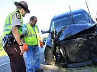 A state trooper and another man look at the front of a crashed vehicle