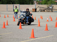 A motorcycle training student navigates through orange cones as an instructor watches