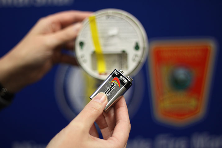 Hands holding a smoke alarm with the battery door open and a 9-volt battery ready to be inserted