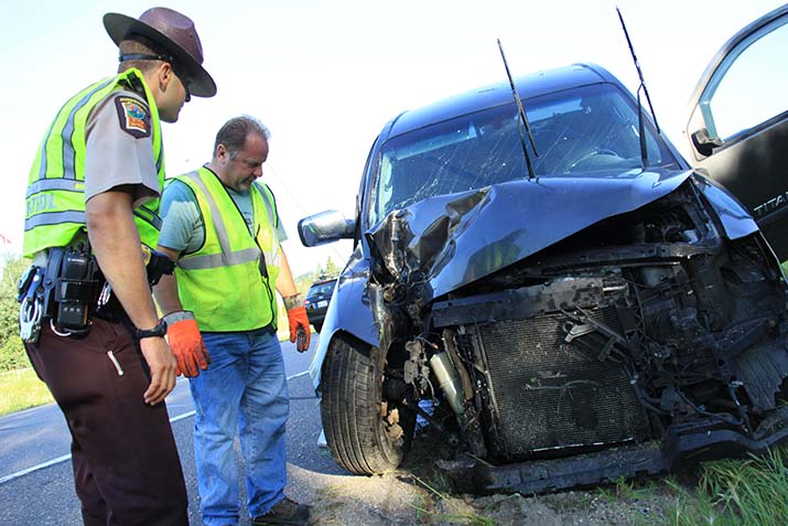 A state trooper and another man look at the front of a crashed vehicle
