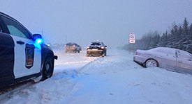 Vehicles on a snow-covered road, a vehicle in the ditch with a State Patrol squad car parked on the shoulder of the road