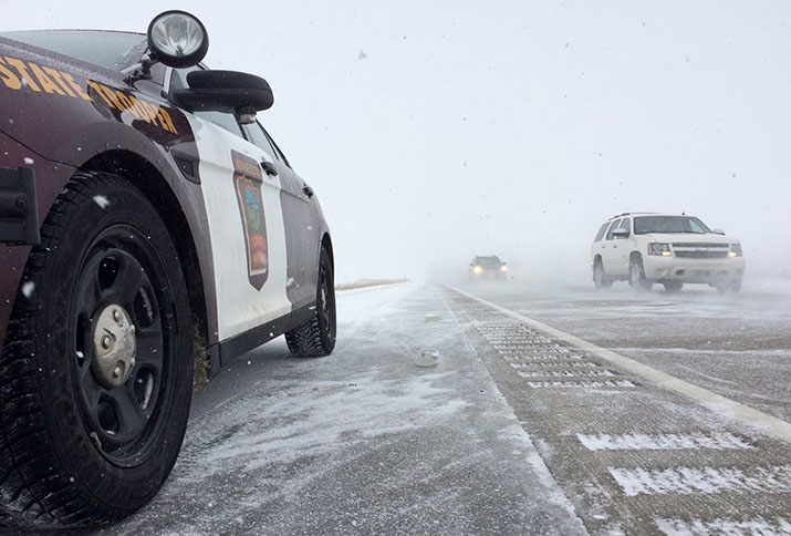 A vehicle driving in blowing snow without its headlights on. A state patrol squad car parked along the shoulder of the road.