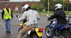 Two riders sitting on motorcycles in front of an instructor in a parking lot during a training session.